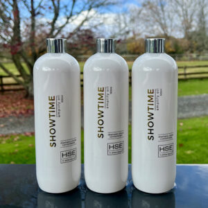Three white bottles labeled "HSE Showtime Coat Oil," containing HSE Hot Oil dramatic shine concentrate from HSE, are displayed on a table with an outdoor background.