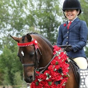 A smiling young rider with braces, wearing formal equestrian attire, sits on a brown horse adorned with a garland of red roses and red ribbons. Trees are visible in the background.