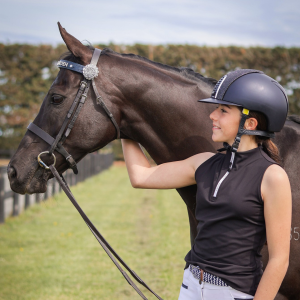 A young woman in a helmet and riding gear stands next to a dark brown horse, holding its reins and smiling while outdoors on a grassy field.