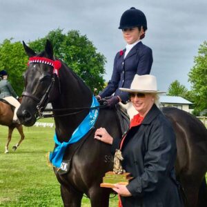 A young equestrian on a black horse with award ribbons poses next to a woman in a hat holding a trophy at an outdoor event.
