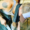 A person wearing a cap and tank top, identified by a visible arm tattoo, is bent over filing a horse's hoof in a workshop with an HSE CoolGroom Hi-Performance Sport Towel hanging from their belt.