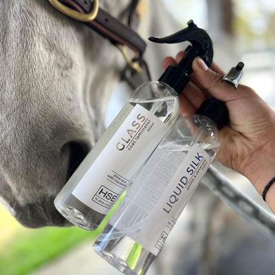 A hand holding two spray bottles labeled "Glass Coat Finishing Spray" and "Liquid Silk." The bottles are positioned in front of a horse's head.