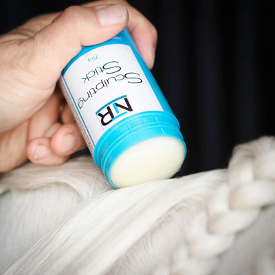 A hand is applying a sculpting stick to white braided hair from a blue container labeled "NIR Sculpting Stick.