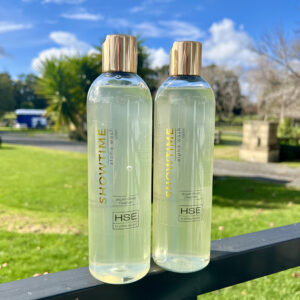 Two bottles of Showtime branded argan oil hair cleanser with gold caps are placed outdoors on a black railing, with grass, trees, and a stone monument in the background. This luxurious HSE Showtime Coat Oil promises to revitalize your hair effortlessly.